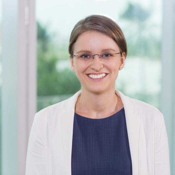 Close up portrait of smiling young businesswoman wearing glasses and looking straight with window and blurry city view outside in background.