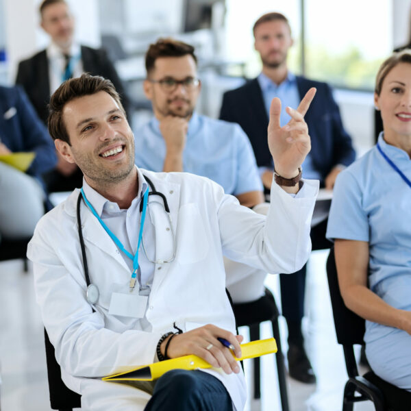 Happy doctor attending a seminar and raising his hand to ask a question at convention center.