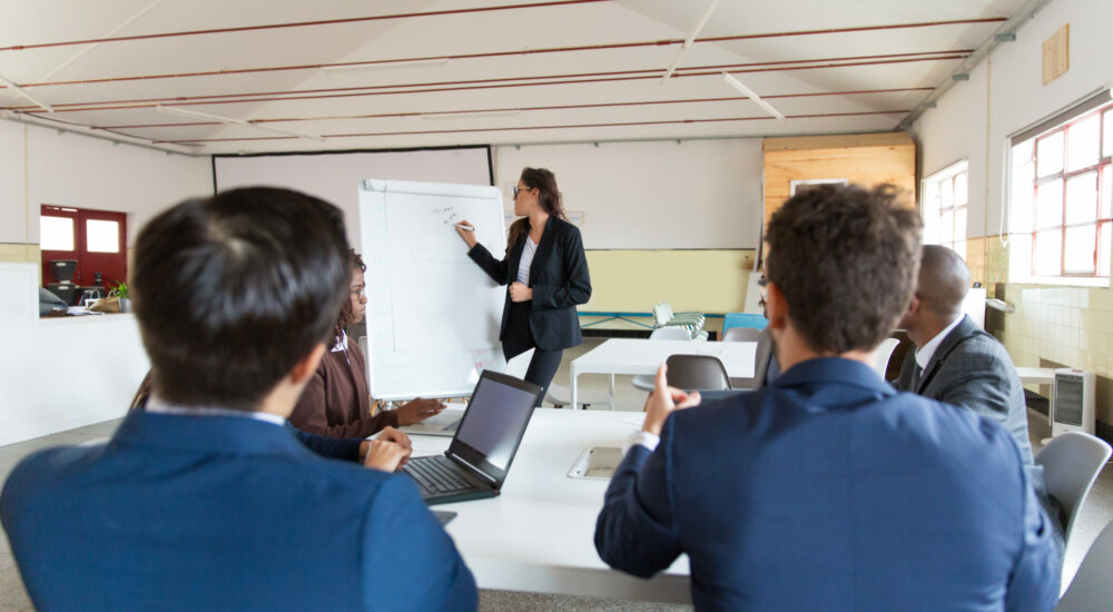 Confident female businesswoman drawing on whiteboard. Group of workers sitting at table and listening speaker. Business meeting concept